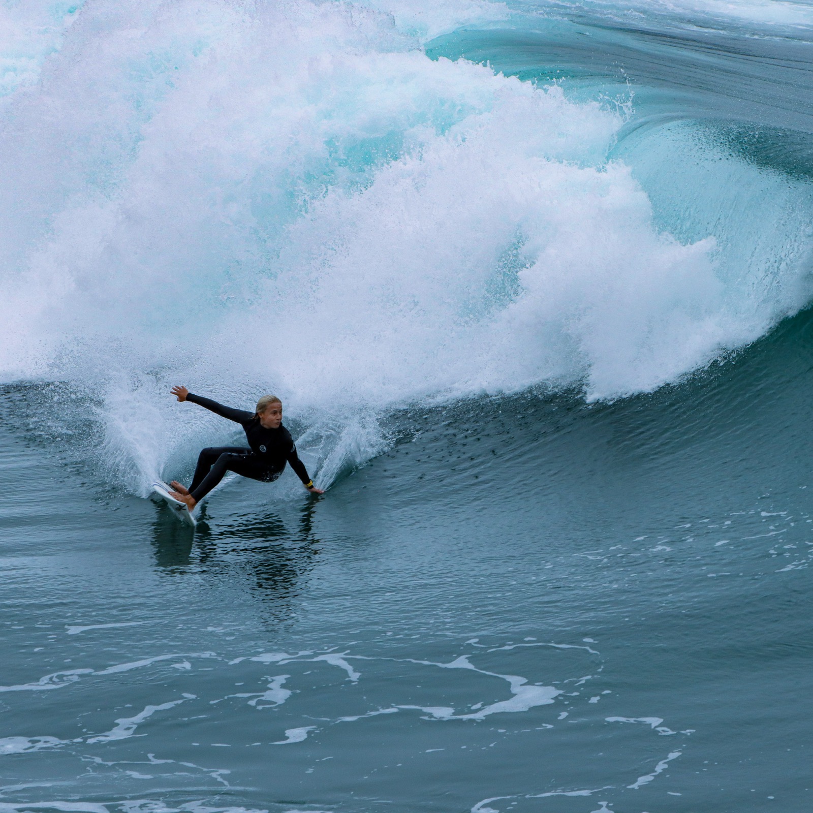 A surfer in a black wetsuit riding low along the face of a large breaking wave. Surf photography by Alice.